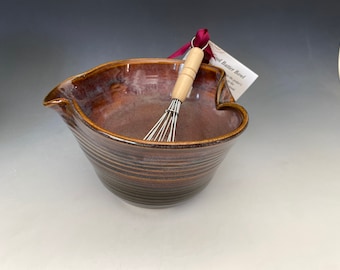May include: A brown ceramic batter bowl with a pouring spout and a wooden-handled whisk. The bowl has horizontal ridges and a tag attached with a maroon ribbon. The bowl is designed for mixing ingredients.