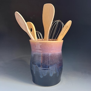 May include: A ceramic utensil holder with a gradient glaze transitioning from a soft pink at the rim to a deep navy blue at the base. The container is filled with various wooden spoons, a wooden spatula, and two metal wire whisks for kitchen use.