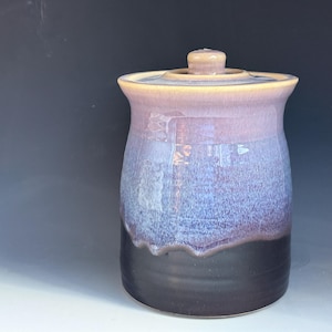 May include: A ceramic canister with a lid, featuring a smooth, glossy glaze that transitions from a light cream color at the top rim to a soft lavender in the middle, and finally to a deep, solid black at the base with a subtle drip effect.