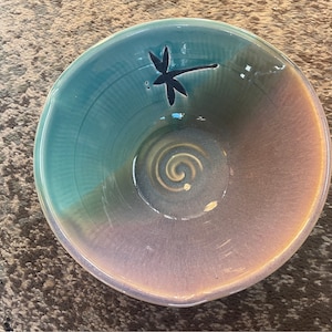 May include: A ceramic bowl with a teal and purple glaze. The bowl has a dragonfly design on the side and a swirl pattern on the bottom.