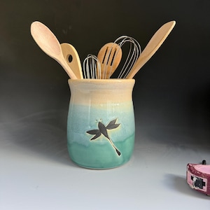 May include: A ceramic utensil holder with a cream and teal ombre glaze featuring a dark dragonfly motif. The holder contains two wooden spoons, a wooden spatula, and two metal whisks. A pink measuring tape is visible in the lower right corner.