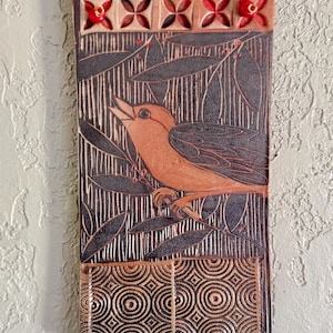May include: A rectangular ceramic wall plaque featuring a carved orange bird perched on a branch amidst dark foliage. The top border displays five carved flower shapes with red centers, while the bottom section shows a repeating circular pattern.