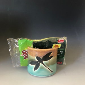 May include: A ceramic cup with a dragonfly design in shades of brown, blue, and green. The cup is sitting on a white surface next to a package of green and yellow sponges.