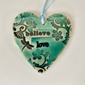 May include: A heart-shaped ceramic ornament in shades of teal and black, with the words "believe love" and a dragonfly design. The ornament has a light blue ribbon for hanging and is decorated with floral and swirl patterns.