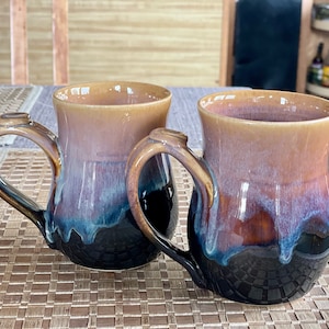 May include: Two ceramic mugs with a brown, purple, and blue glaze. The mugs have a unique, rounded shape and a handle on each side.