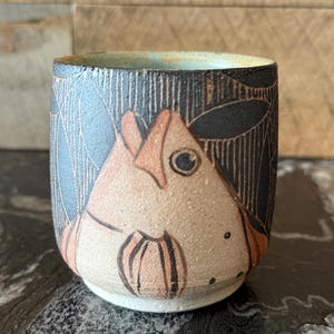 May include: A small ceramic cup with a black and white design of a fish. The fish is painted in shades of orange and brown.