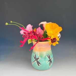 May include: A small, hand-painted ceramic vase with a turquoise base and orange-pink top, adorned with black dragonfly silhouettes. The vase holds a vibrant bouquet of colorful flowers, including yellow, pink, and red blooms, creating a cheerful display.