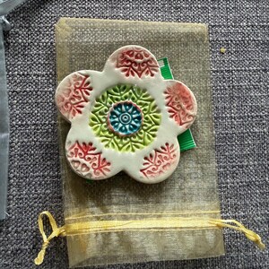 May include: A white ceramic flower-shaped ornament with red and green accents. The center features a blue and green design. It rests on a small, beige fabric pouch with a yellow ribbon tie, suggesting a gift or decorative item.