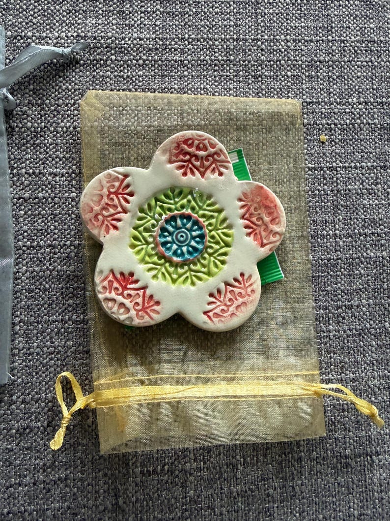 May include: A white ceramic flower-shaped ornament with red and green accents. The center features a blue and green design. It rests on a small, beige fabric pouch with a yellow ribbon tie, suggesting a gift or decorative item.