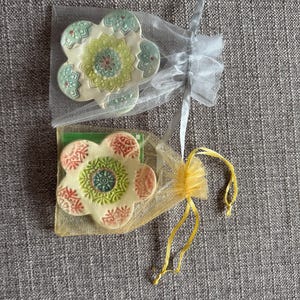 May include: Two flower-shaped ceramic coasters, each in a sheer organza bag. One coaster has a green, yellow, and red floral design. The other has a pink, green, and blue floral design. The bags are tied with ribbons.