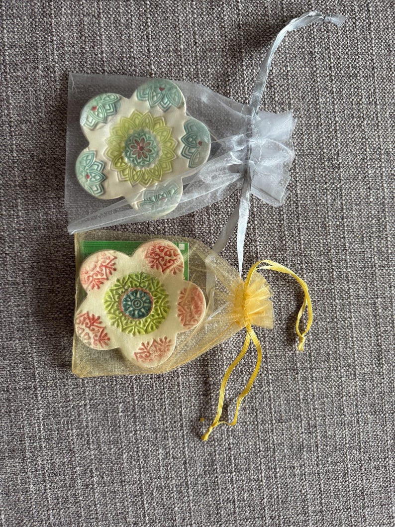 May include: Two flower-shaped ceramic coasters, each in a sheer organza bag. One coaster has a green, yellow, and red floral design. The other has a pink, green, and blue floral design. The bags are tied with ribbons.