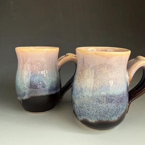 May include: Two ceramic mugs featuring a reactive glaze with a color gradient transitioning from a light cream at the rim to a soft lavender, then a mottled blue, and finally a deep dark brown at the base, each with a curved handle.