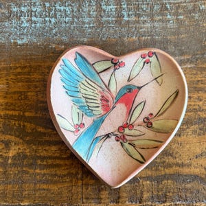 May include: Heart-shaped ceramic dish with a hand-painted hummingbird design. The bird features blue wings, a red throat, and a long beak, perched on a branch with berries. The dish has a pinkish hue and is set against a wooden background.