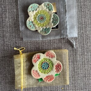 May include: Two ceramic flower-shaped coasters. One is in a sheer gray bag, the other is on a tan paper-like material tied with a yellow ribbon. The coasters have a white base with colorful floral designs in green, red, and blue.
