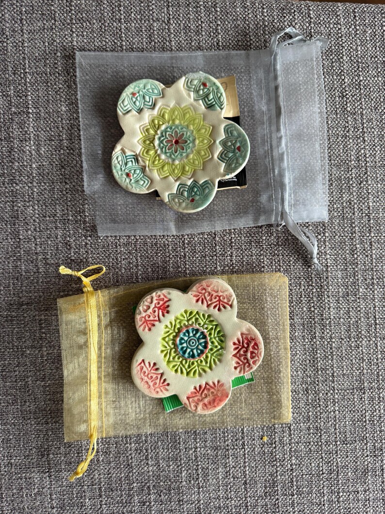 May include: Two ceramic flower-shaped coasters. One is in a sheer gray bag, the other is on a tan paper-like material tied with a yellow ribbon. The coasters have a white base with colorful floral designs in green, red, and blue.