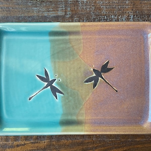 May include: A rectangular ceramic tray with a teal glaze on one side and a brown glaze on the other. The tray has two dragonfly designs, one on each side.