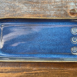 May include: A blue ceramic serving tray with a brown rim and three decorative swirls on the right side.