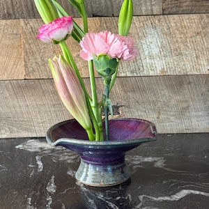 May include: A ceramic flower vase with a deep purple interior and a gray exterior holds a floral arrangement. The arrangement includes pink carnations, a pink and white ranunculus, and green lily buds. The vase sits on a black marble surface.