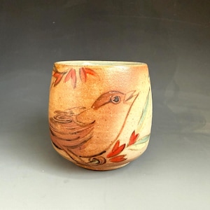 May include: A rounded ceramic cup with a tan, speckled glaze featuring a hand-painted brown bird perched among stylized red leaves and green foliage. The vessel has a smooth, curved silhouette and a wide opening, showcasing a natural, earthy aesthetic with visible brushstrokes throughout the painted design.