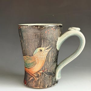 May include: A ceramic mug with a dark, textured charcoal exterior featuring a hand-painted illustration of a small bird with orange and green plumage perched on a branch. The mug has a light blue, curved handle and a vertical etched line pattern.
