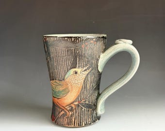 May include: A ceramic mug with a dark, textured charcoal exterior featuring a hand-painted illustration of a small bird with orange and green plumage perched on a branch. The mug has a light blue, curved handle and a vertical etched line pattern.
