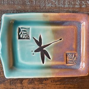 May include: A rectangular ceramic tray featuring a gradient glaze transitioning from turquoise to warm brown. The center displays a dark dragonfly silhouette, flanked by two small square indentations, each containing a relief of a leafy branch.