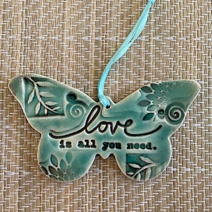 May include: A teal blue ceramic butterfly ornament with the words "love is all you need." hanging from a light blue ribbon.