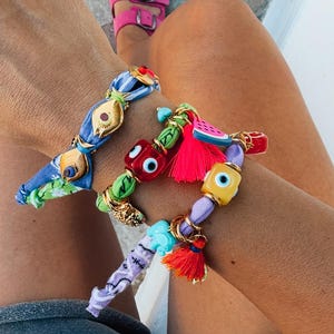 May include: A person wearing three colorful beaded bracelets with gold accents. The bracelets feature various beads, including evil eye charms, tassels, and fabric-wrapped elements. The person is also wearing pink sandals.