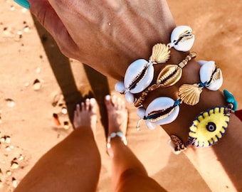 May include: A hand wearing several bracelets with white seashells, gold accents, and a blue and yellow evil eye charm. The bracelets are on a tan background.
