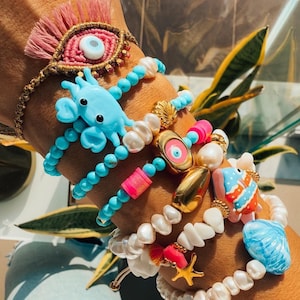 Puede incluir: Una pila de pulseras coloridas con varios encantos, incluyendo un cangrejo azul, un mal de ojo dorado, un pez rosa y blanco y una concha azul. Las pulseras están hechas de cuentas turquesa, blancas y rosas.