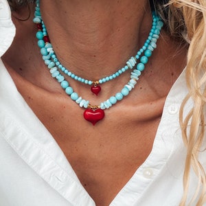 May include: A layered necklace with turquoise and white beads and a red heart pendant. The necklace is worn by a person with blonde hair and a white shirt.