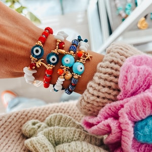 Op de afbeelding: Een stapel kleurrijke armbanden met blauwe boze oogcharms, witte kralen en rode en blauwe kralen. De armbanden zitten om de pols van een persoon.