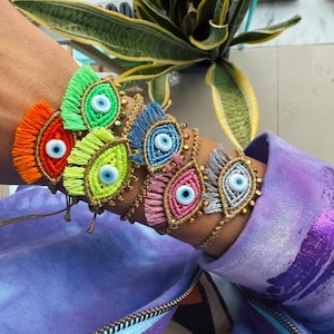 Peut inclure: Un ensemble de bracelets colorés avec des charmes de mauvais œil. Chaque bracelet est fait avec une couleur de fil différente et a un accent de perle dorée. Les bracelets sont empilés sur le poignet d'une personne.