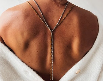 May include: A back view of a person wearing a white sweater with a long, double-strand necklace. The necklace is made of two chains, one gold and one silver, that are connected at the back of the neck and hang down the back.