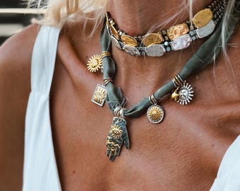 May include: A layered necklace set featuring a choker with alternating gold and silver hammered oval links and a sage green fabric ribbon necklace adorned with multiple metal charms including suns, a rectangular eye, and a hand pendant.