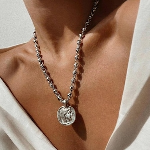 Könnte beinhalten: Eine silberfarbene Halskette mit einem Anhänger, der das Profil einer Frau zeigt. Die Halskette hat eine Perlenkette. Der Anhänger ist kreisförmig und zeigt ein detailliertes Relief des Gesichts der Frau. Die Halskette wird gegen ein weißes Hemd getragen.