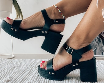 May include: Black platform sandals with a buckle closure and silver studs. The sandals have a thick wooden platform and heel.