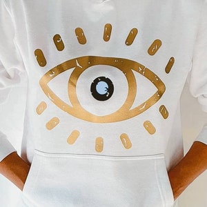May include: White hooded sweatshirt with a gold evil eye graphic design. The eye has a blue pupil and is surrounded by gold rays.
