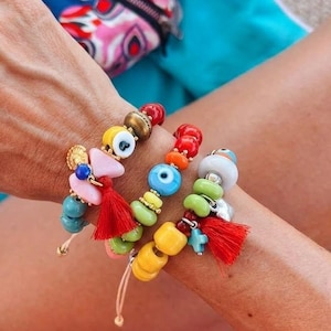 Può includere: Un set di tre braccialetti di perline con elementi in ceramica e plastica nei toni del giallo, verde, blu e rosso. I braccialetti includono piccoli ciondoli a forma di occhio, una perla rosa a forma di farfalla e due piccole nappe rosse.