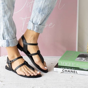 May include: A pair of black leather sandals with a buckle strap and a t-strap design. The sandals are worn by a person wearing blue jeans.