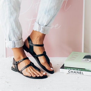 May include: A pair of black leather thong sandals with a buckle closure. The sandals are worn by a person wearing light blue jeans.
