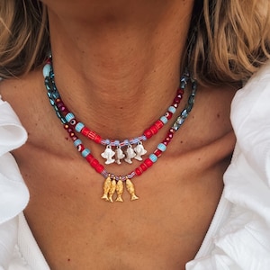 以下が含まれることがあります： 赤、青、白のビーズでできた重ね付けネックレス。ネックレスには、小さな銀色の魚のチャームと、下からぶら下がっている金色の魚のチャームが付いています。