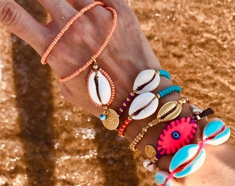May include: A hand wearing a variety of bracelets with colorful beads, seashells, and a pink evil eye charm. The bracelets are stacked on the wrist and are a mix of different styles and colors.