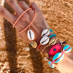 Op de afbeelding: Een hand met verschillende armbanden met gekleurde kralen, schelpen en een roze boze oog-bedel. De armbanden zijn gestapeld op de pols en zijn een mix van verschillende stijlen en kleuren.