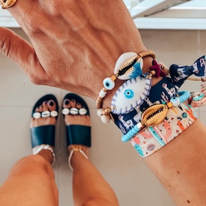 Op de afbeelding: Een hand met blauwe en witte nagellak met een oogontwerp op de nagels. De hand draagt een gouden ring met een schelpontwerp. De hand draagt ook verschillende armbanden met schelp- en boze oogcharms. De armbanden zitten op een pols met een kleurrijk, gepatroneerd stoffen armband.