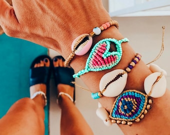 May include: A hand with light blue nail polish with an evil eye design on one finger. The hand is wearing four bracelets with seashells and colorful beads. The bracelets feature a pink heart, a blue eye, and two white seashells.