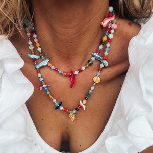 Op de afbeelding: Een gelaagde ketting met kleurrijke kralen, schelpen en bedels. De ketting heeft een verscheidenheid aan kleuren, waaronder rood, blauw, groen en geel. De bedels omvatten een schelp, een ster en een kleine rode peper.