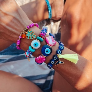 Peut inclure: Un ensemble de bracelets colorés avec des charmes de mauvais œil et un charme en feuille d'or. Les bracelets sont au poignet d'une personne.