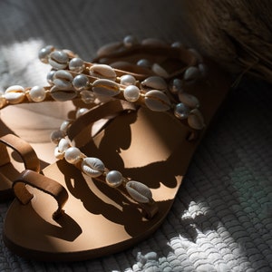 May include: A pair of brown leather sandals with white shell and pearl embellishments. The sandals have a simple design with a single strap across the toe and a strap around the ankle.