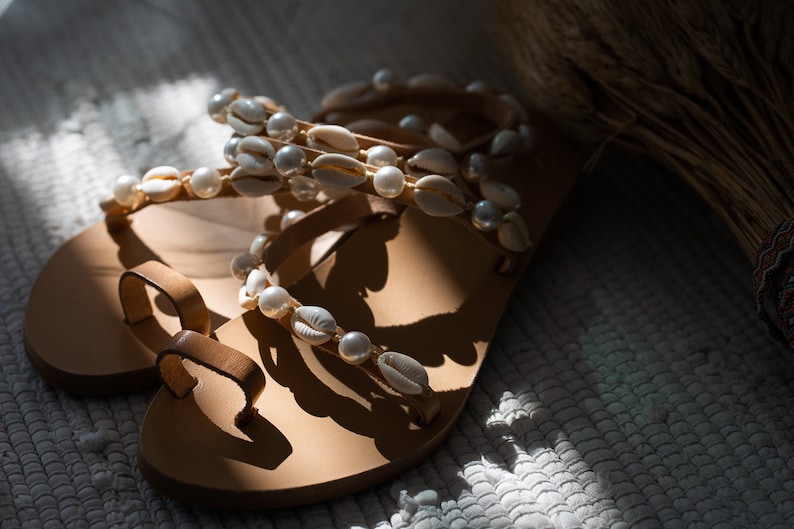 May include: A pair of brown leather sandals with white shell and pearl embellishments. The sandals have a simple design with a single strap across the toe and a strap around the ankle.